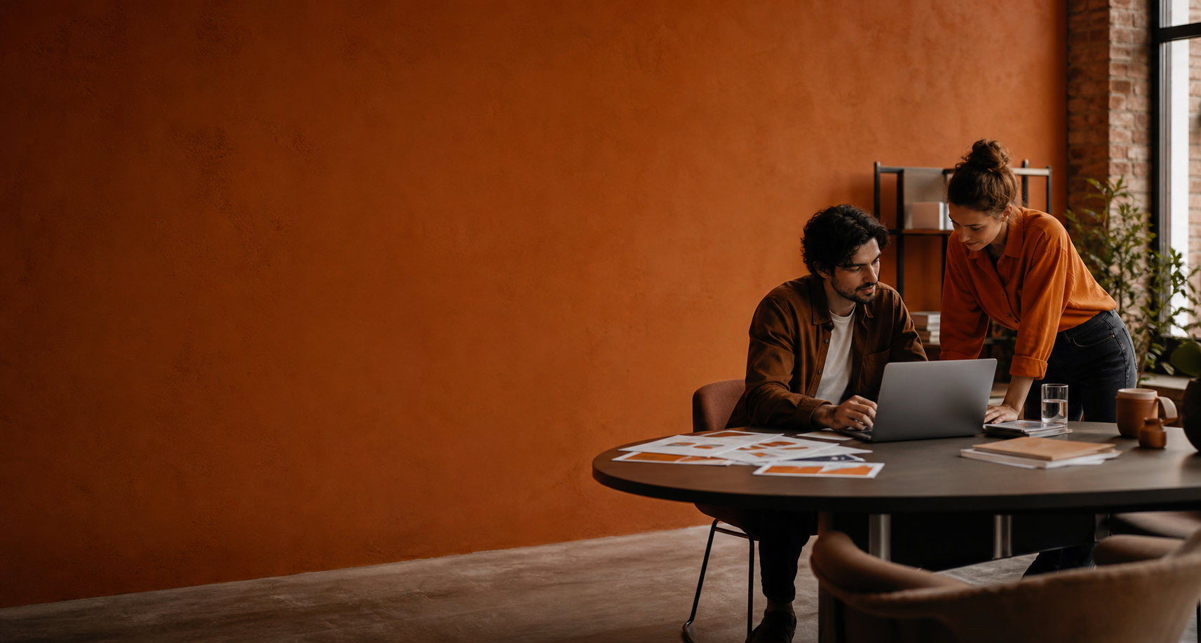 Deux collègues discutent autour d’un ordinateur portable dans un bureau au mur orange